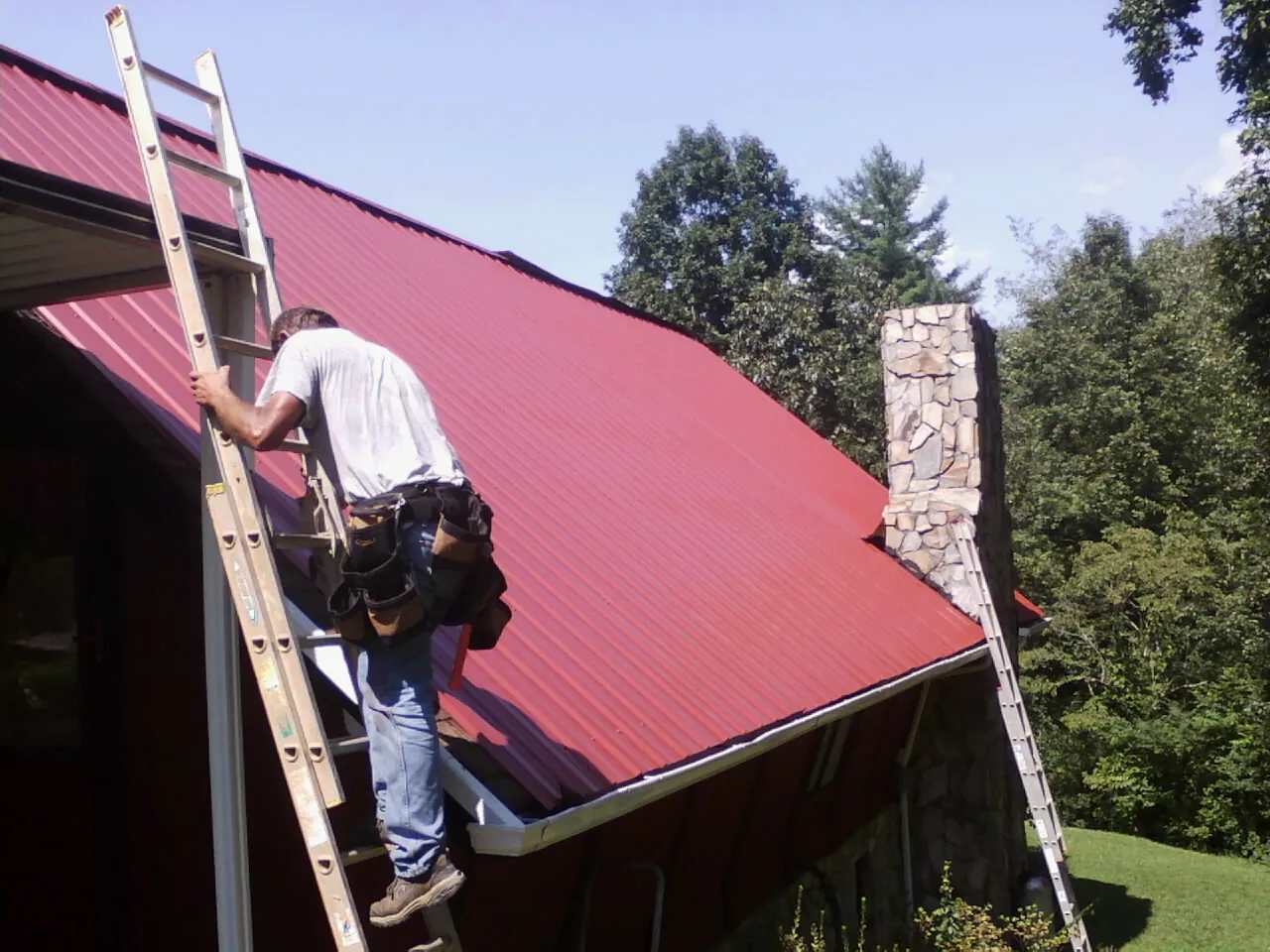 Roofer on a ladder beside a red metal roof and stone chimney — Soffit & Fascia Repair by Roofing Brothers in Gales Creek